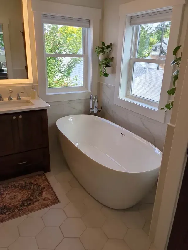 Bathroom with a white freestanding tub, windows, and plants. Light-colored tile floor, dark wood vanity.