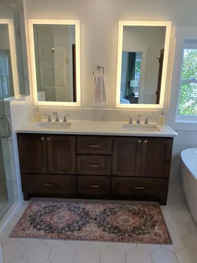 Bathroom with double vanity, two lighted mirrors, dark wood cabinet, marble countertop, and patterned rug.
