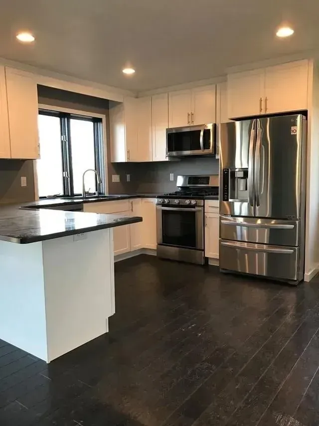 Modern white kitchen with stainless steel appliances, dark wood floors, and granite countertops.