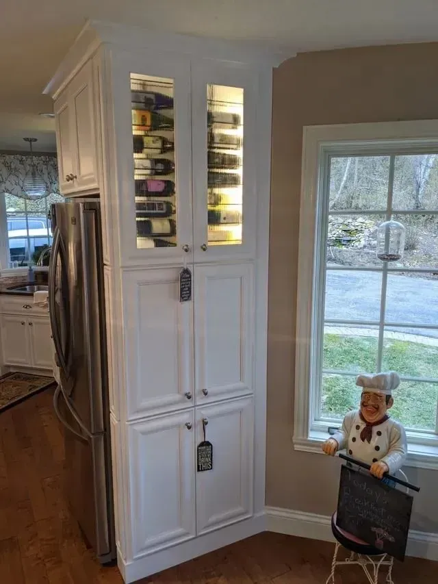 White cabinet with wine rack, stainless fridge, and chef statue by a window.