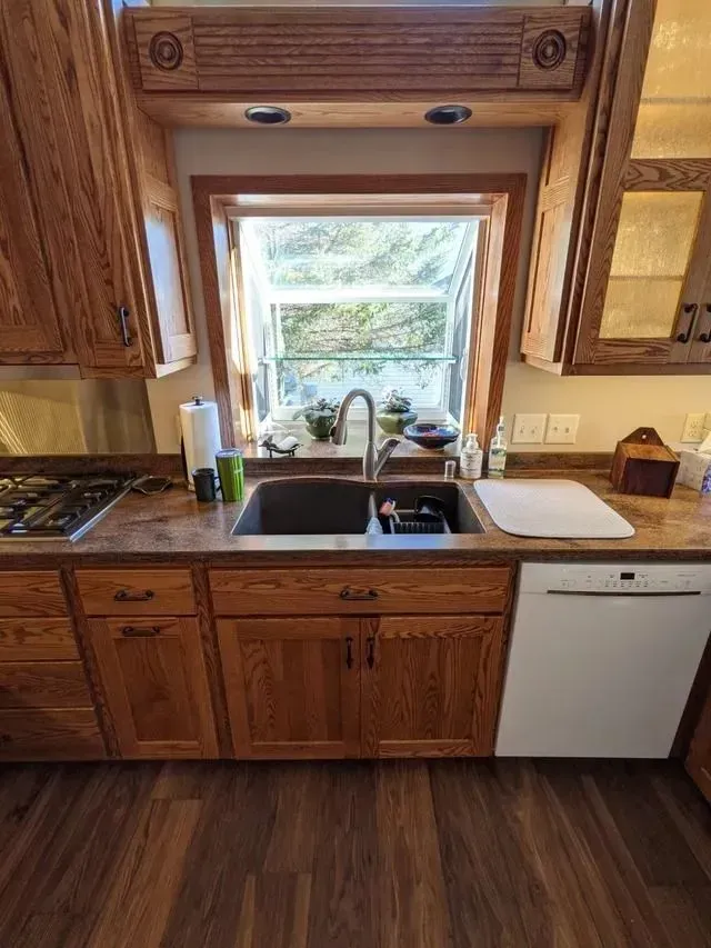 Kitchen with oak cabinets, sink, and window overlooking trees. Brown countertops and dark wood flooring.
