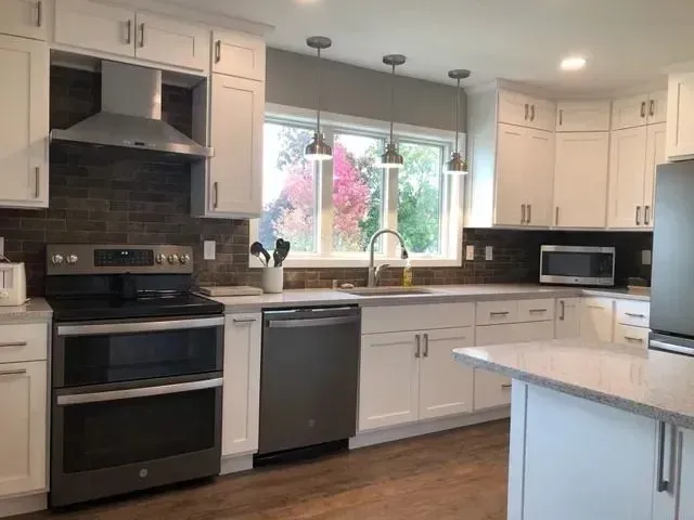 White kitchen with stainless steel appliances, grey countertops, and a window with colorful foliage.