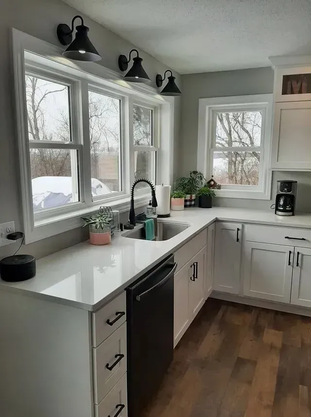 Bright white kitchen with a window over the sink, black faucet and lights, and dark wood floors.
