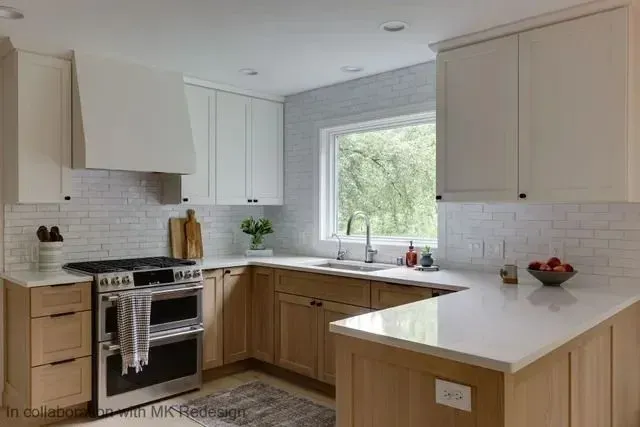 Modern kitchen with white and wood cabinets, white countertops, and a window overlooking greenery.