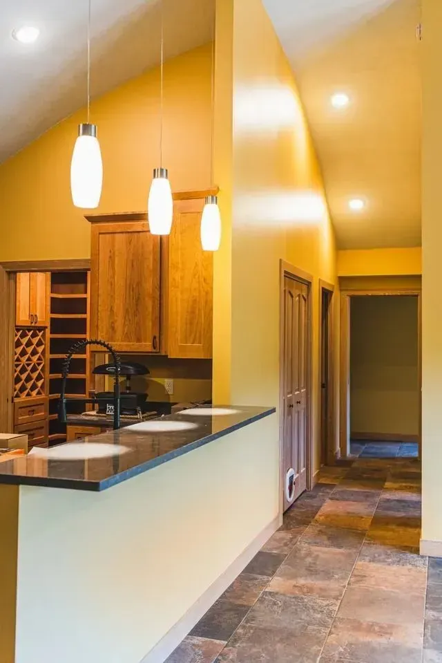 Yellow kitchen with hanging lights, wooden cabinets, and a black countertop, leading to a hallway.