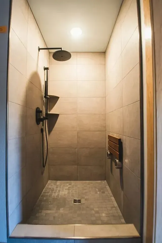 Walk-in shower with beige tile walls, stone floor, showerhead, and a fold-down wooden seat.