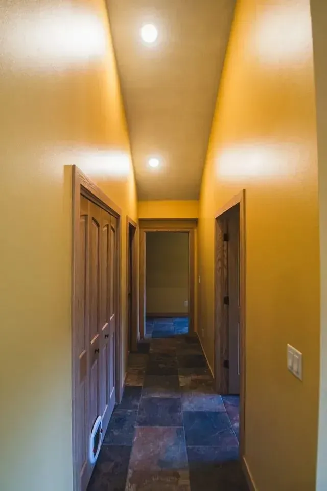 Narrow hallway with yellow walls, dark tile floor, wooden doors, and recessed ceiling lights.