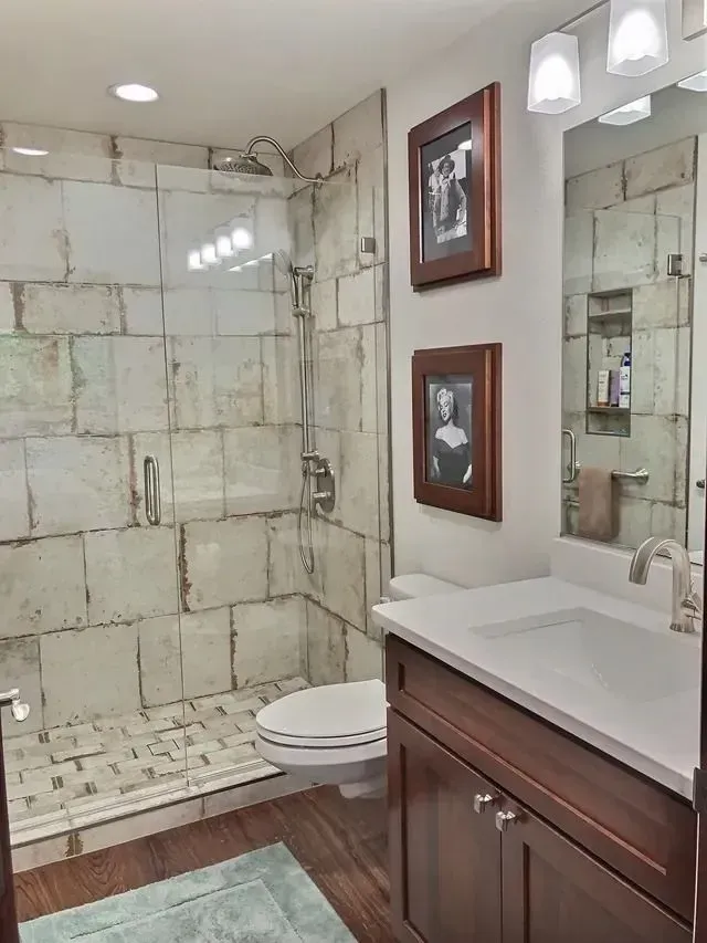 Bathroom with tiled shower, vanity, toilet. Brown cabinets, white countertops, wooden floor, framed photos on wall.