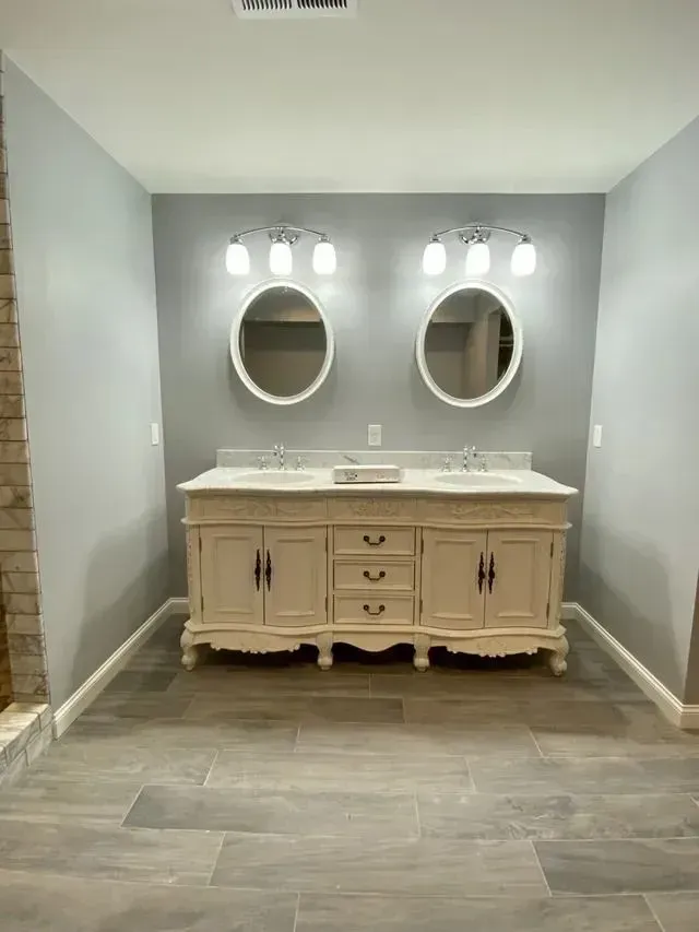 Bathroom with a double vanity in cream color, two oval mirrors, and gray walls and floor.