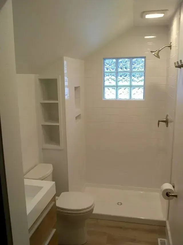 Bathroom with white tile walls, shower, toilet, and shelving, glass block window.