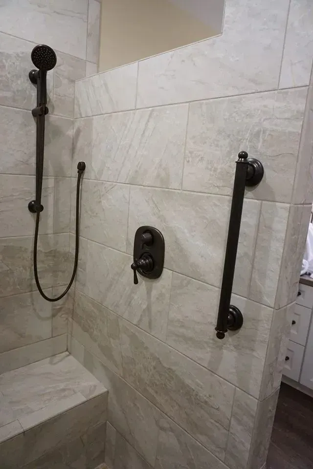 Shower with dark fixtures, including a hand shower and grab bar, on light-colored tile walls.
