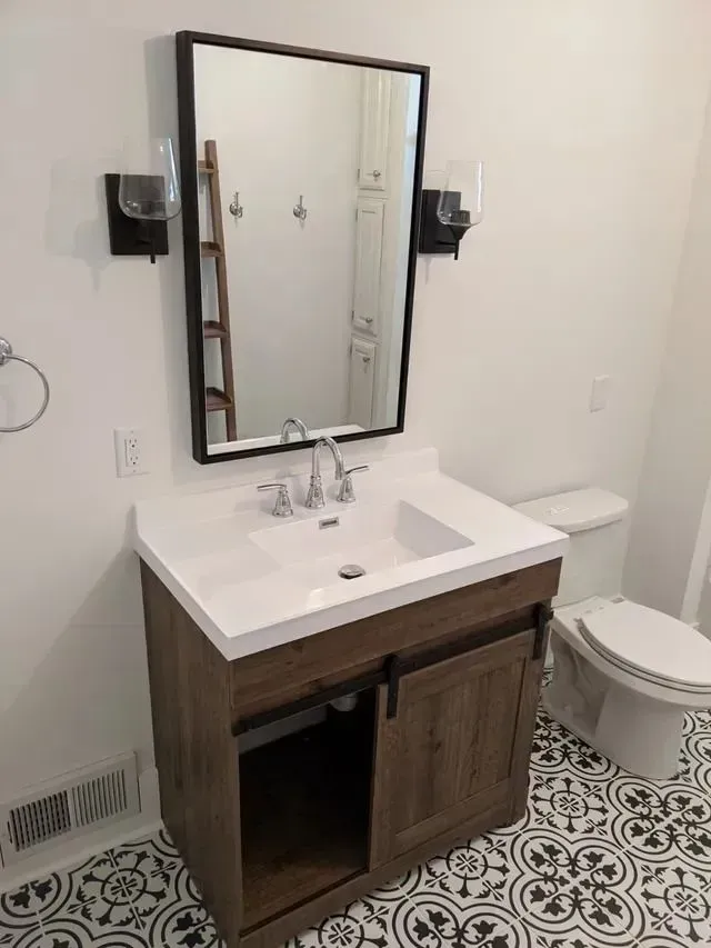 Bathroom vanity with mirror, sconces, and sliding cabinet doors. White countertop, wood cabinet, patterned floor.