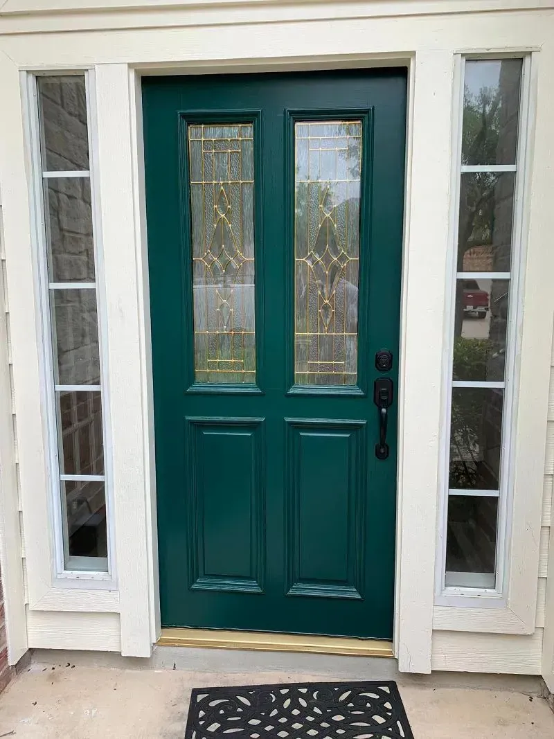 Green front door with glass panels, flanked by windows, black doormat, white trim.