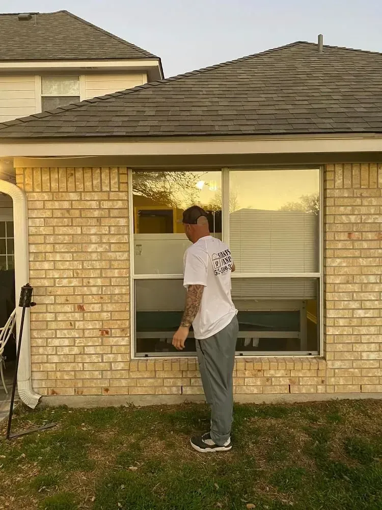 Man cleaning a window outside a brick house, wearing a white t-shirt and grey pants.