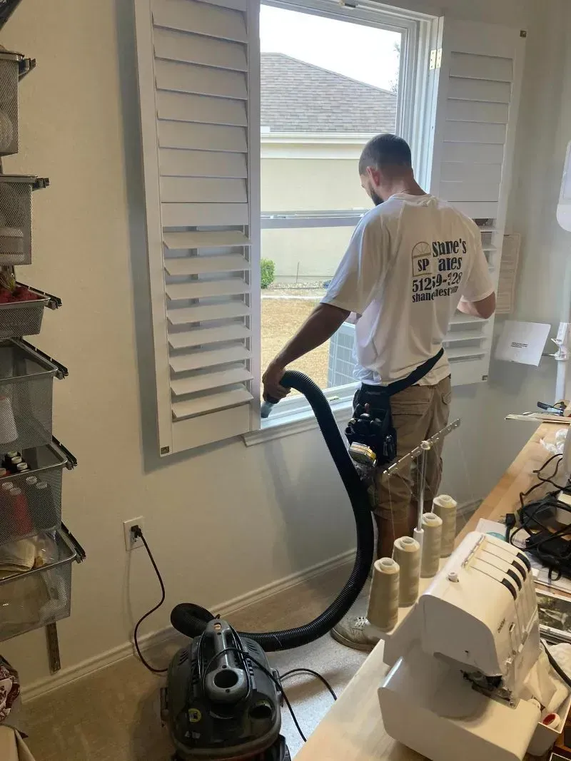 A person vacuums window shutters indoors. White shutters frame a window; a sewing machine sits nearby.