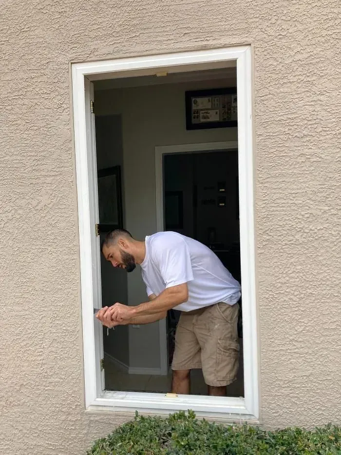 Man working on a window frame, indoors. Exterior wall is tan. He wears shorts, white shirt.