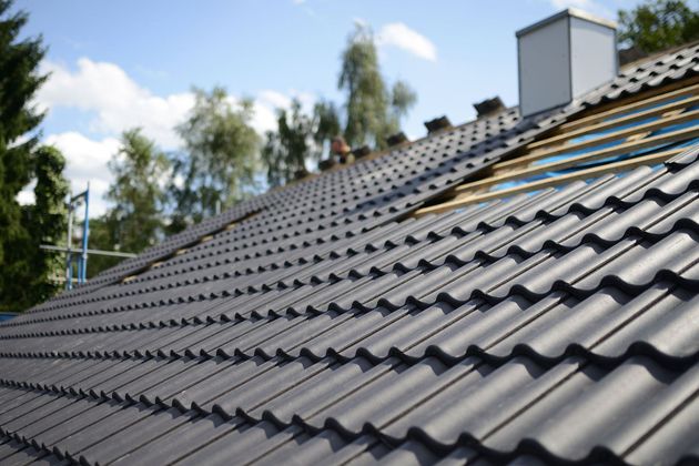Gray tiled roof under repair with exposed underlayment and chimney against a blue sky