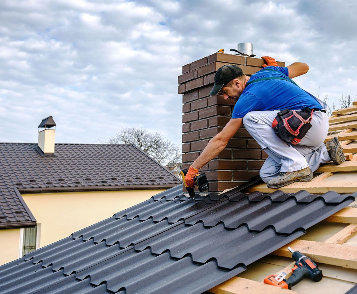 Worker installing roof tiles on a house, kneeling beside a brick chimney with tools nearby.