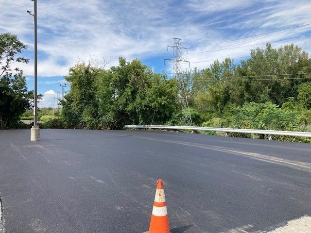 Newly paved asphalt road with trees and power lines in the background, a safety cone in foreground.
