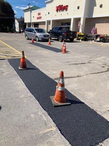 Fresh asphalt strip in a parking lot, marked with orange cones, in front of a store.