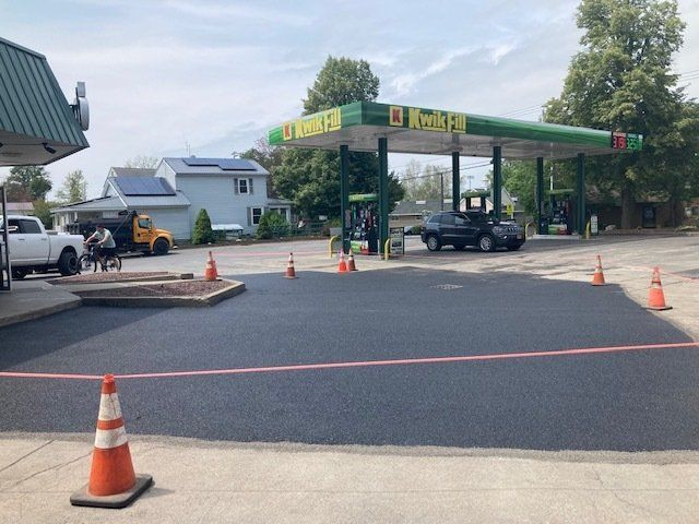 Gas station asphalt repair in progress. Freshly paved black surface, orange cones, pumps, and a vehicle are present.