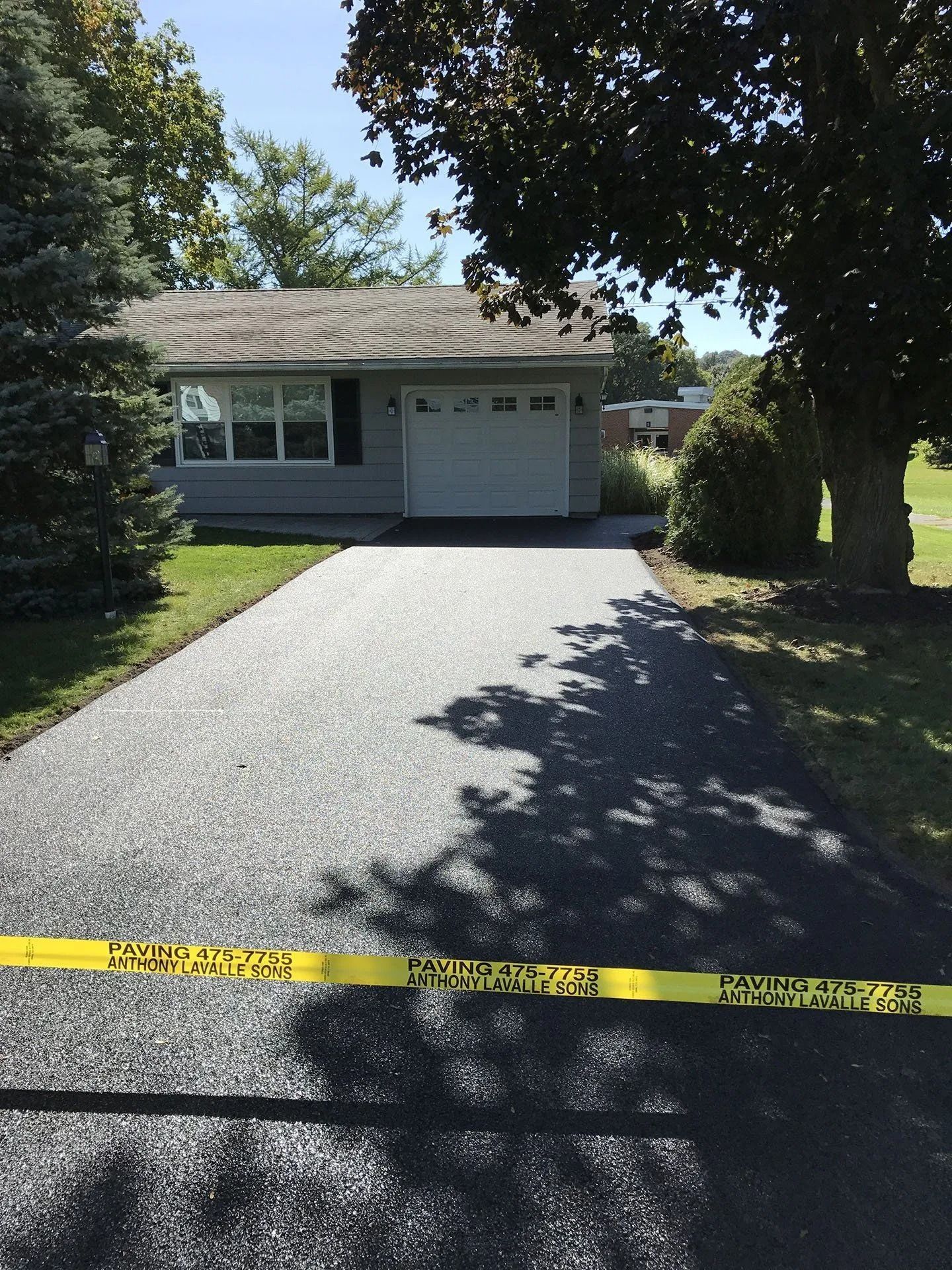 Dark asphalt driveway leading to a one-story house with a two-car garage. Yellow caution tape is across the driveway.