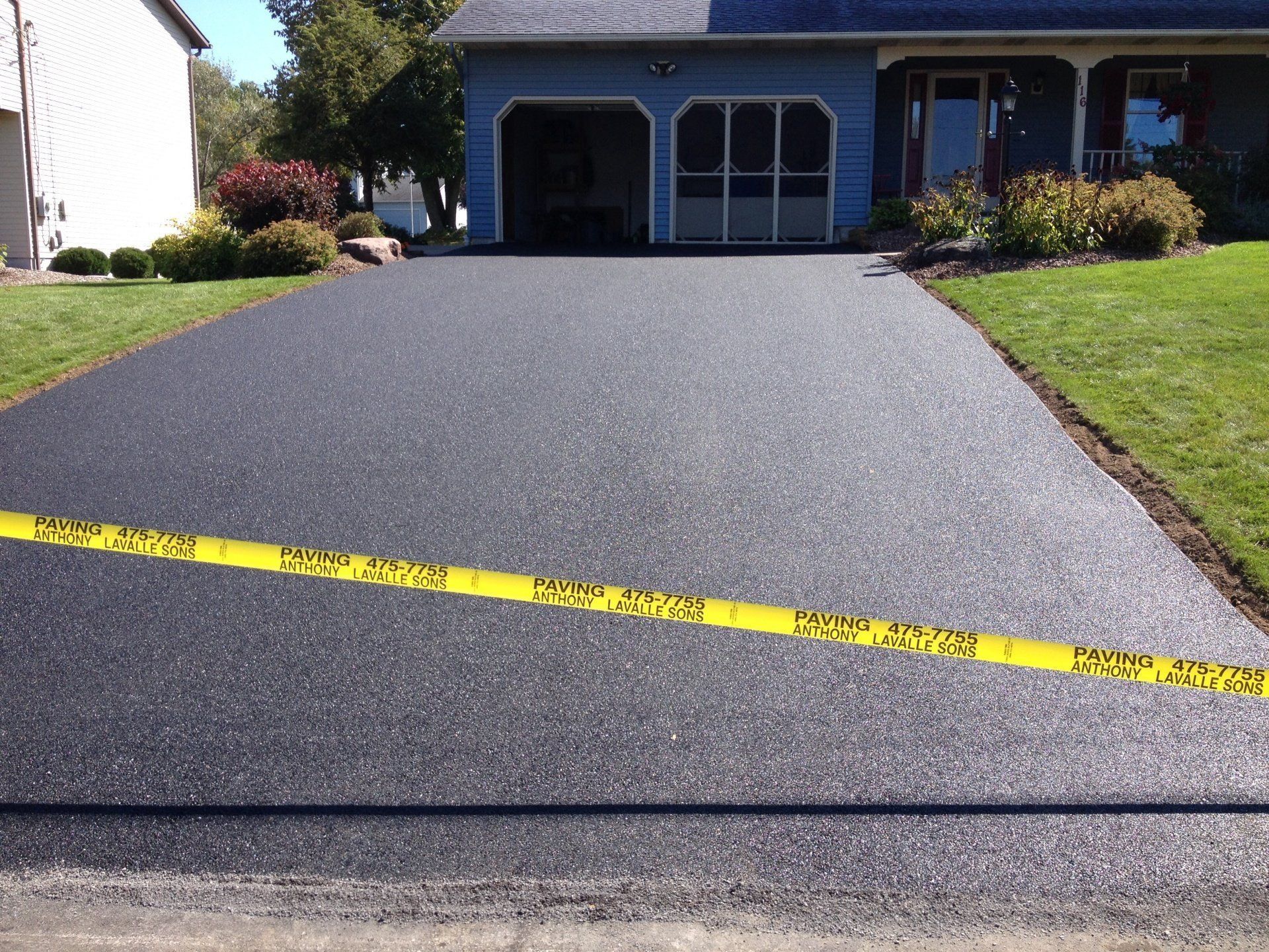 Newly paved black asphalt driveway in front of a blue house with a garage and green lawn, marked with yellow caution tape.