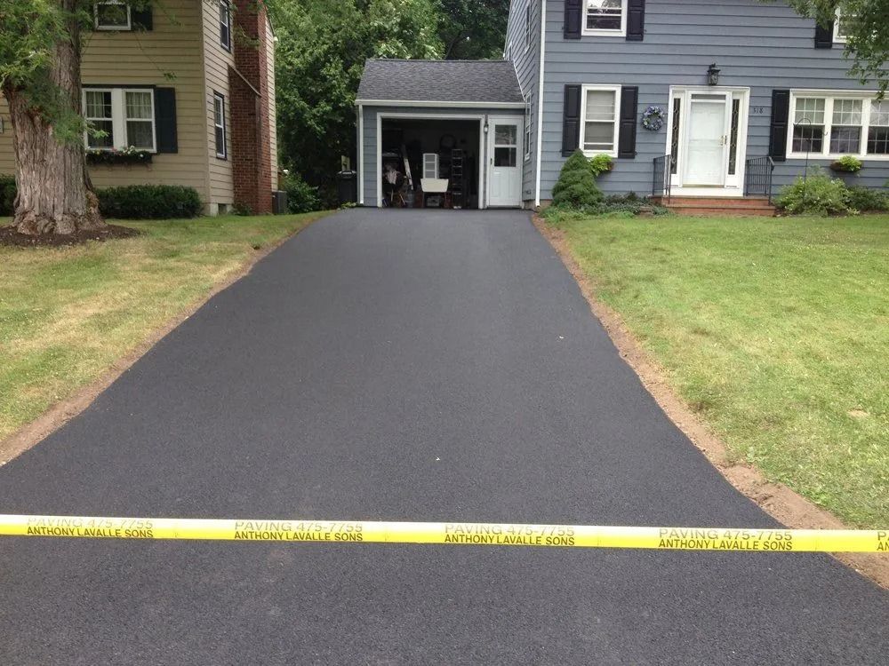 Newly paved asphalt driveway leading to a garage, blocked by yellow caution tape, flanked by grass. Houses in the background.