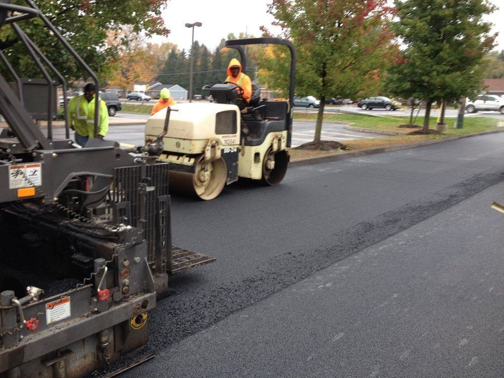 Asphalt paving in progress: worker operates roller, another watches, trees in the background.