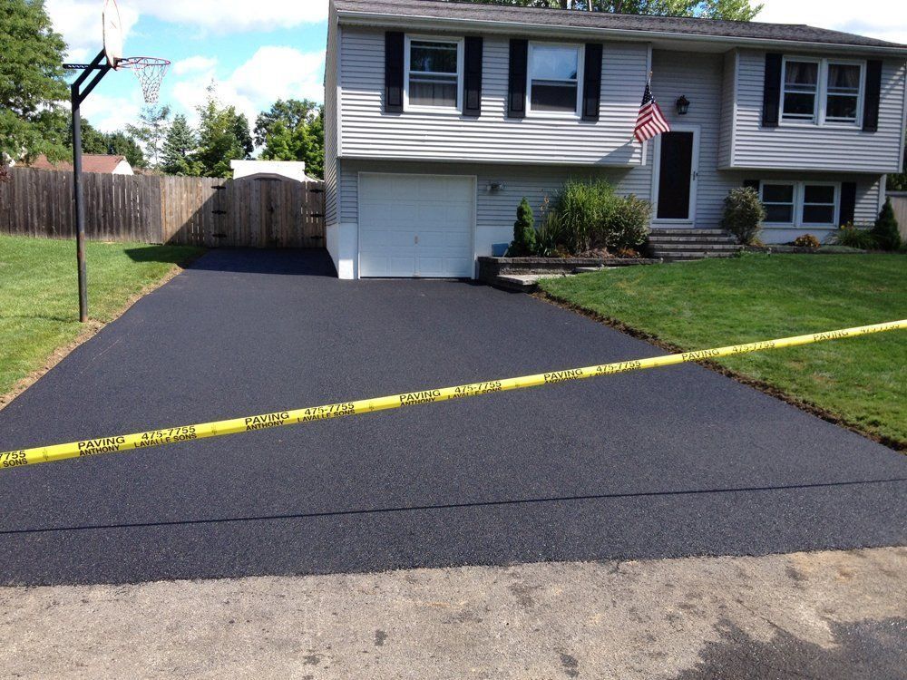 Freshly paved black asphalt driveway in front of a gray house, caution tape, and a basketball hoop.