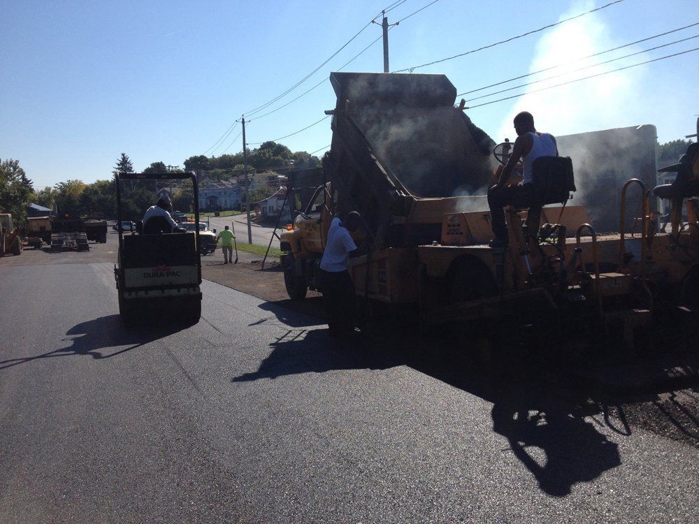Road paving in progress; asphalt paver, roller, and workers on street.