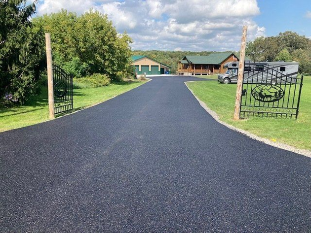 Black asphalt driveway leading to a house with decorative black gates. Green lawn, blue sky, and trees.