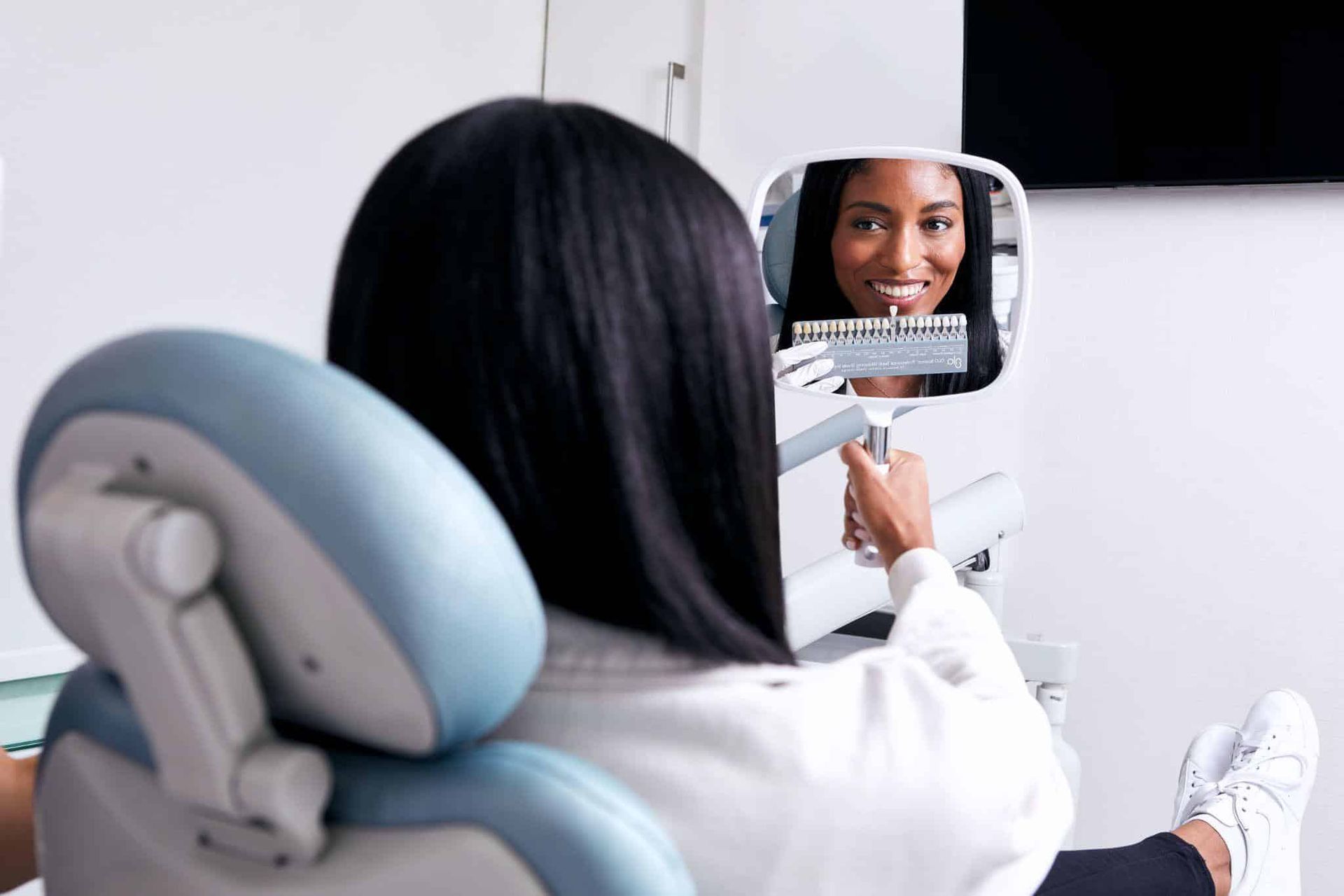 A woman is sitting in a dental chair looking at her teeth in a mirror.
