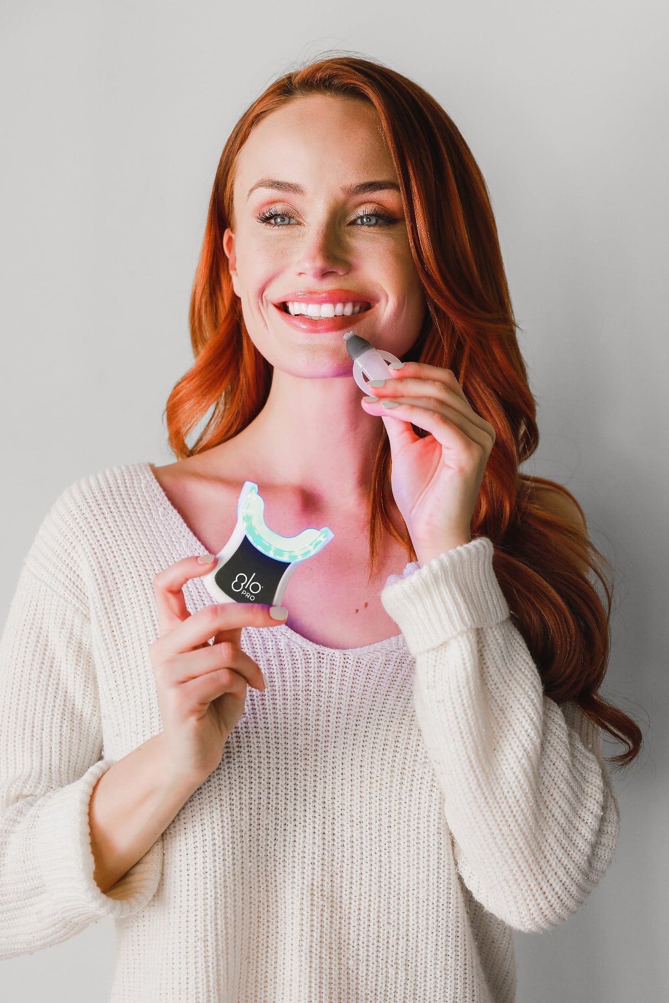 A woman in a white sweater is holding a device to whiten her teeth.