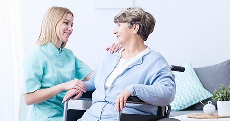 A caregiver in teal scrubs smiles at an older woman in a wheelchair, indoor setting.