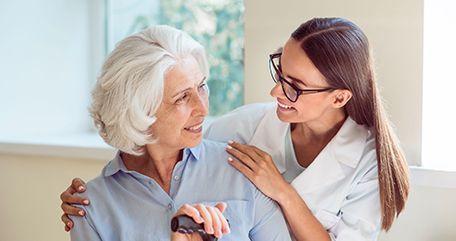 Woman with gray hair, holding a cane, smiles at a healthcare provider who has her arm around her.