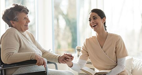 Woman in wheelchair and caregiver laughing together, holding hands, near a window.