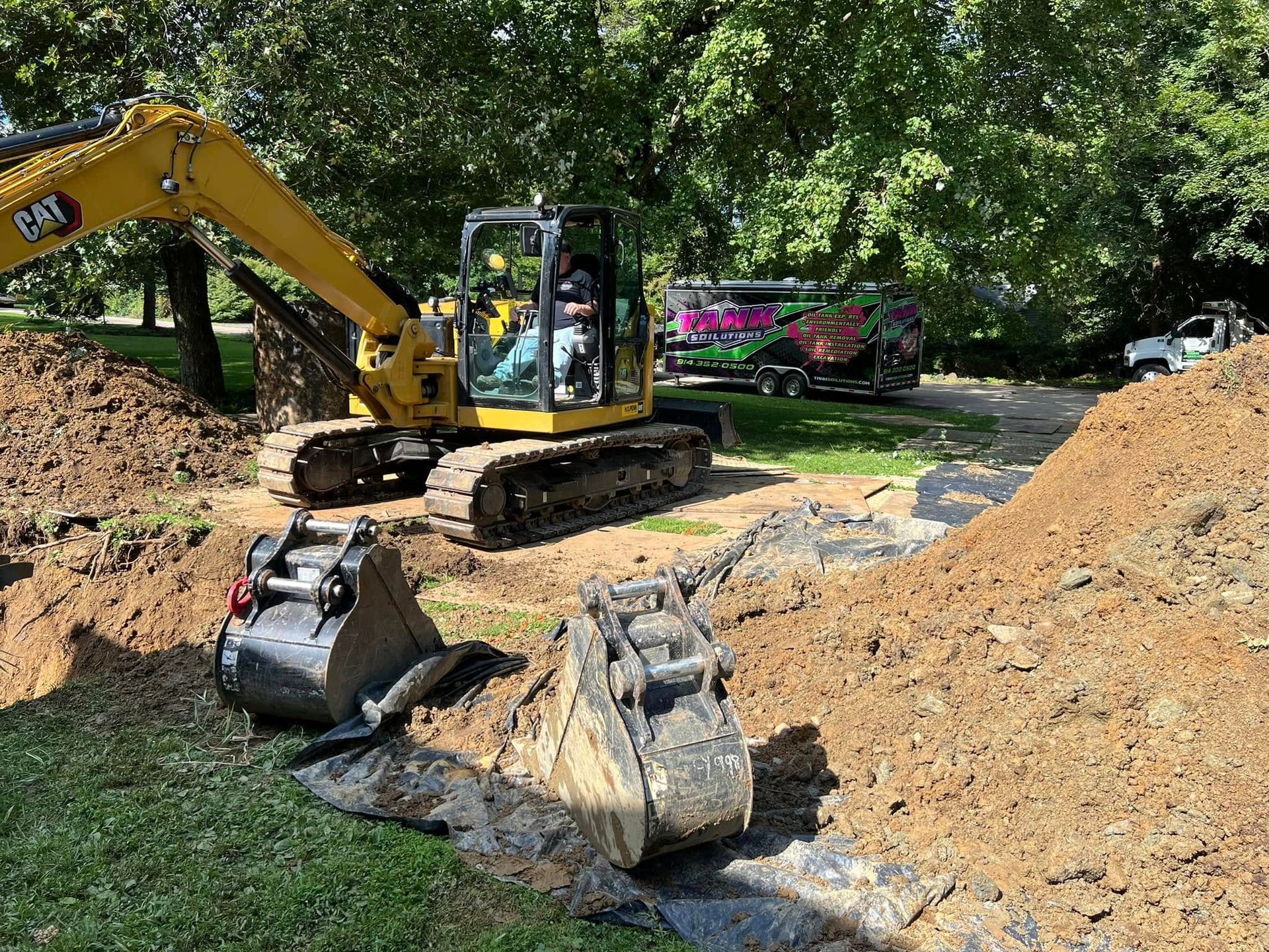 A yellow CAT excavator sits on a construction site with two detached buckets in the foreground and a trailer nearby.