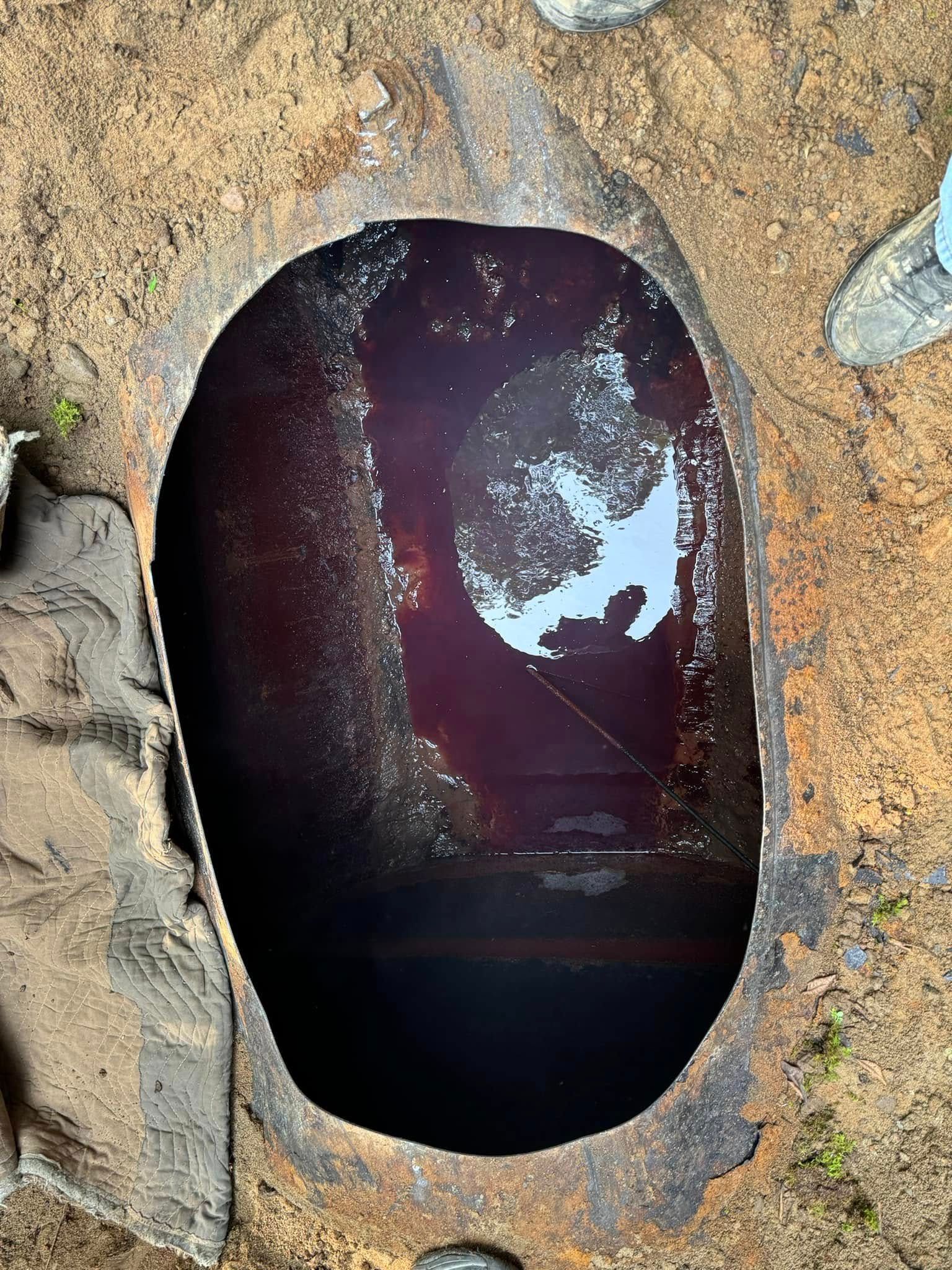 A circular hole in the ground reveals a dark, reddish-purple liquid inside a rusty container.