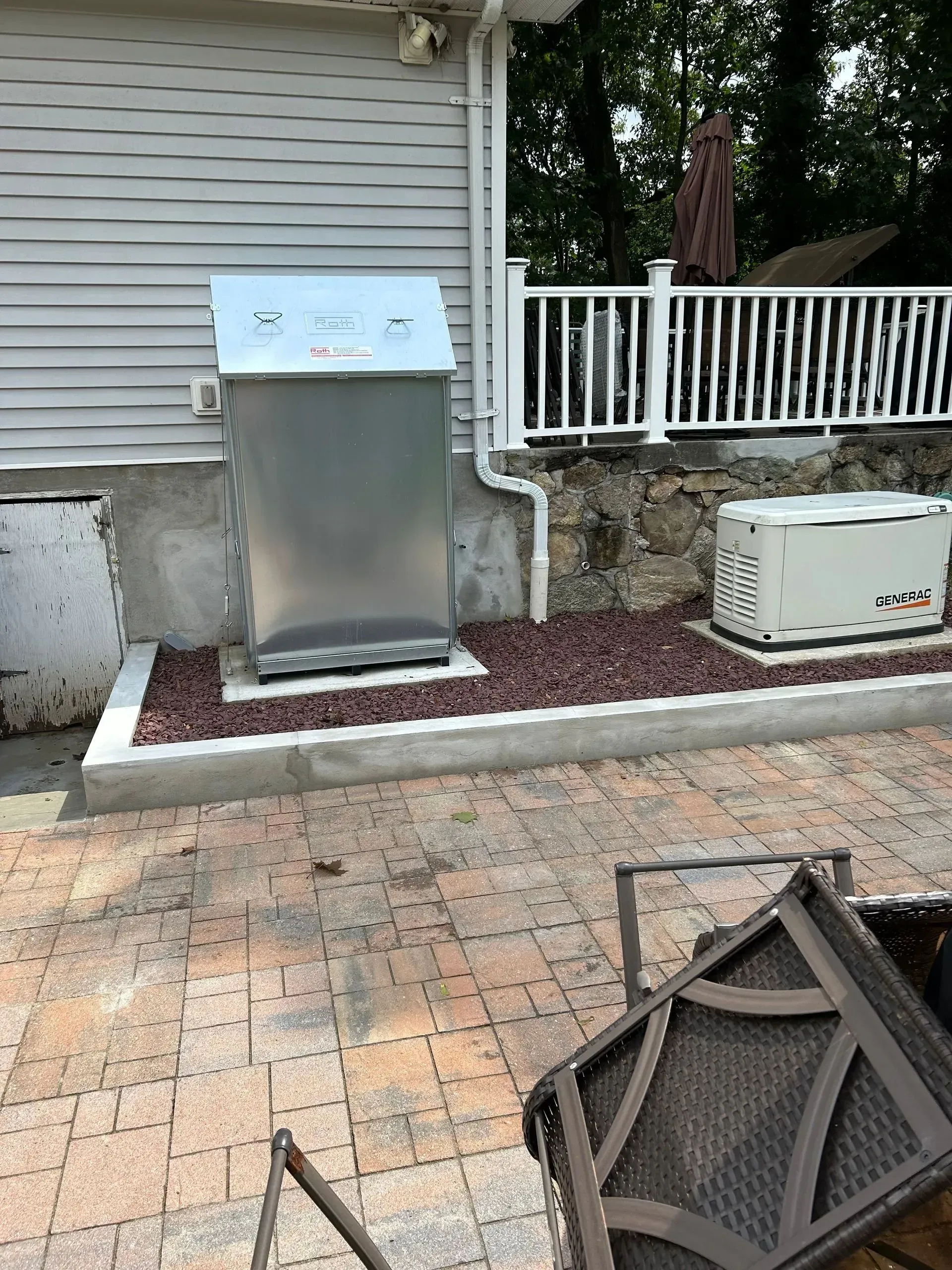 A metal utility enclosure and a white backup generator sitting on a stone-bordered gravel pad next to a patio.