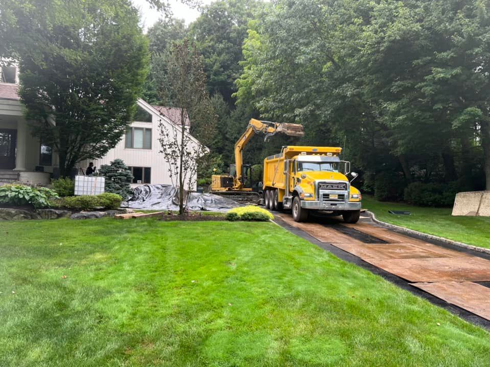 A yellow dump truck and excavator work in the driveway of a house surrounded by trees and a green lawn.