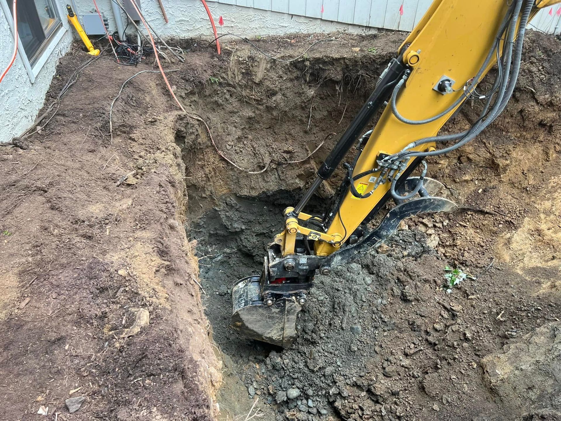 A yellow excavator digging a trench in the dirt next to the side of a house.