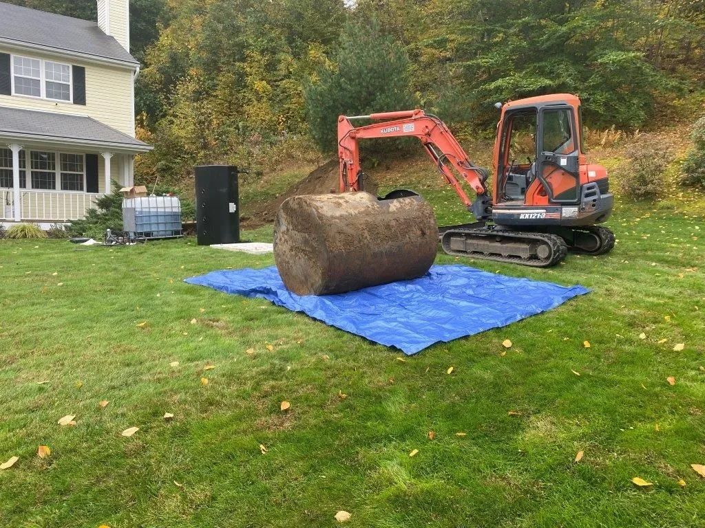 An orange excavator sits in a grassy yard next to a large, excavated cylindrical tank resting on a blue tarp.