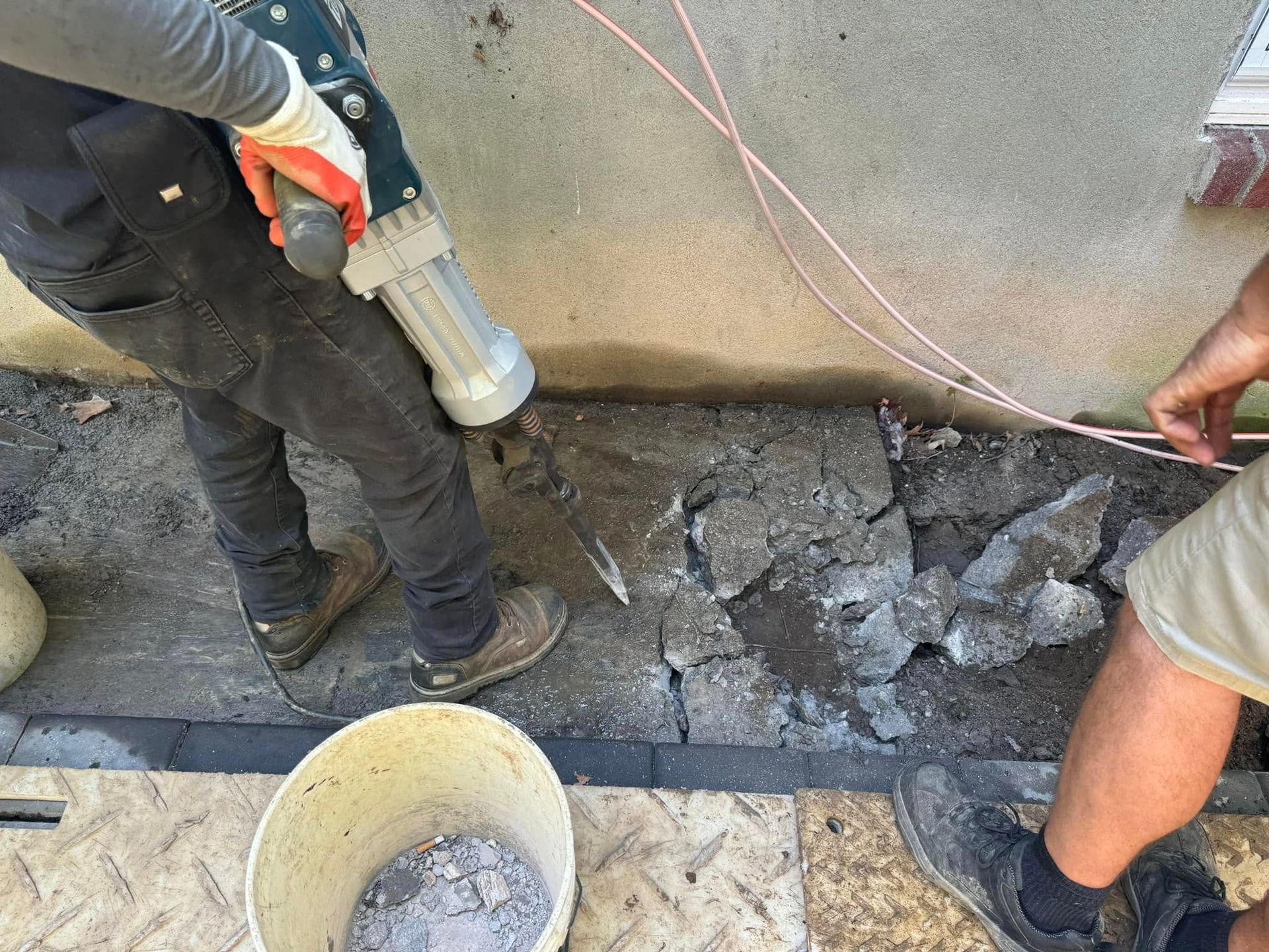 A worker uses an electric jackhammer to break up concrete next to a wall, with a bucket nearby and someone standing by.