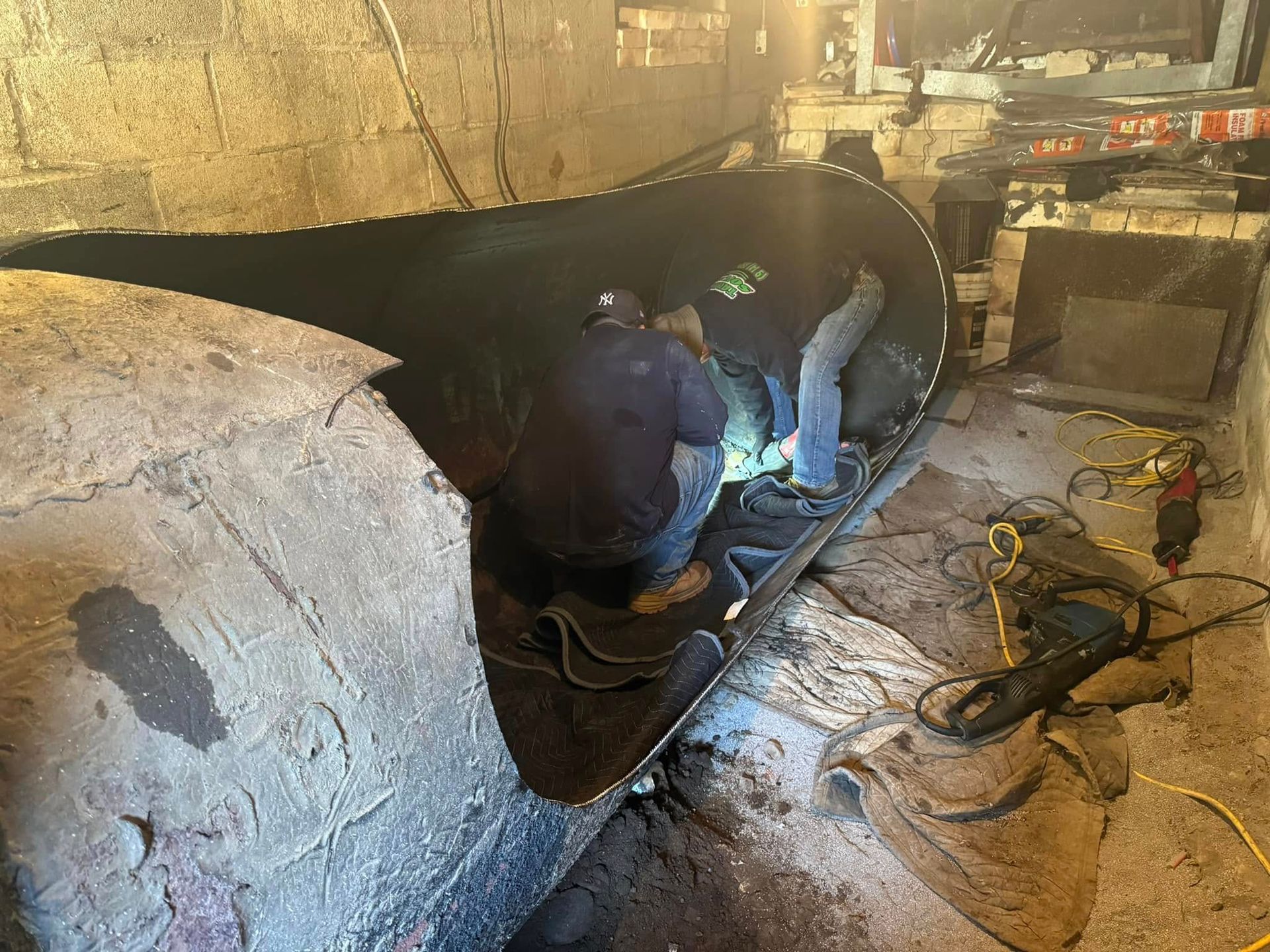 Two workers in dark clothing repair a large, horizontal metal tank in a dimly lit, unfinished workspace.