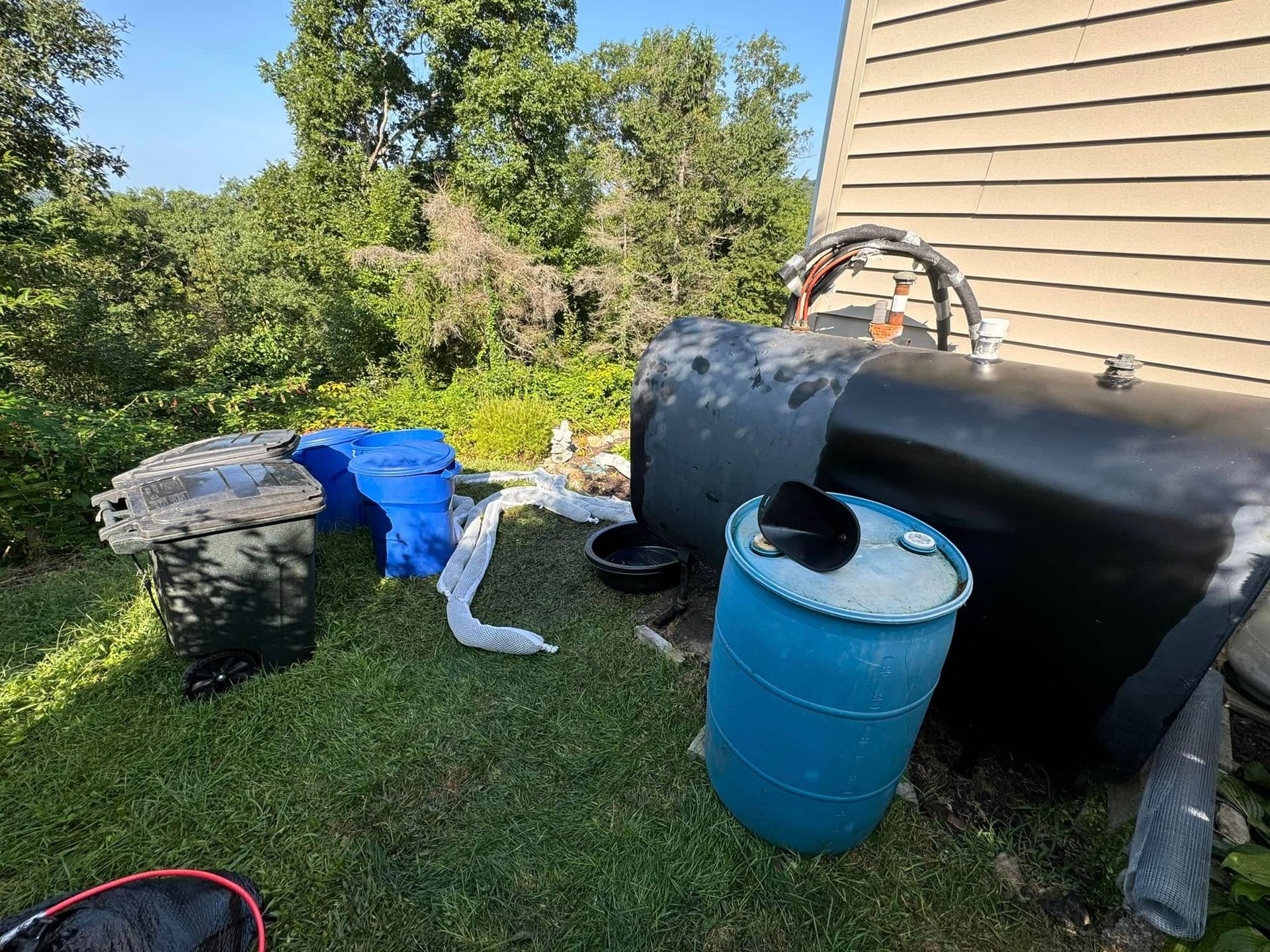An outdoor scene featuring a large black fuel tank, a blue barrel, and plastic containers in a grassy, wooded yard.