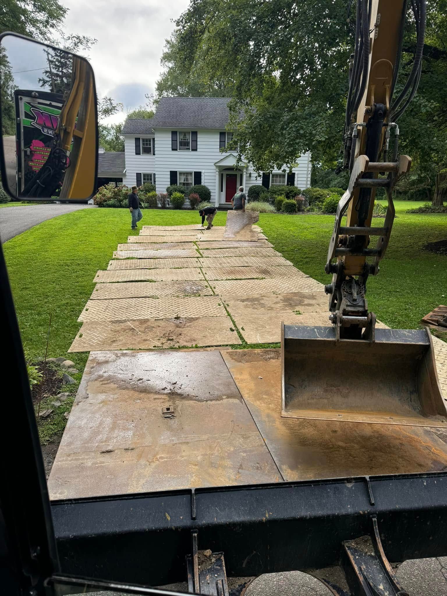 An excavator sits on heavy steel plates laid across a lawn in front of a white two-story house.