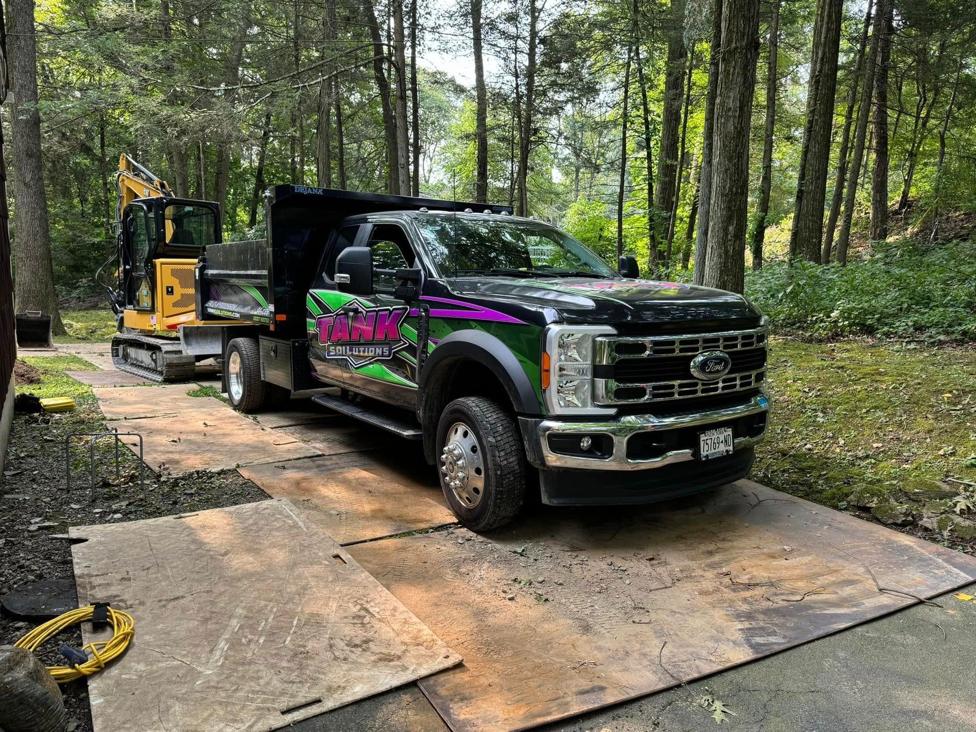 A black dump truck with purple and green graphics sits on plywood sheets in a wooded area with an excavator behind it.