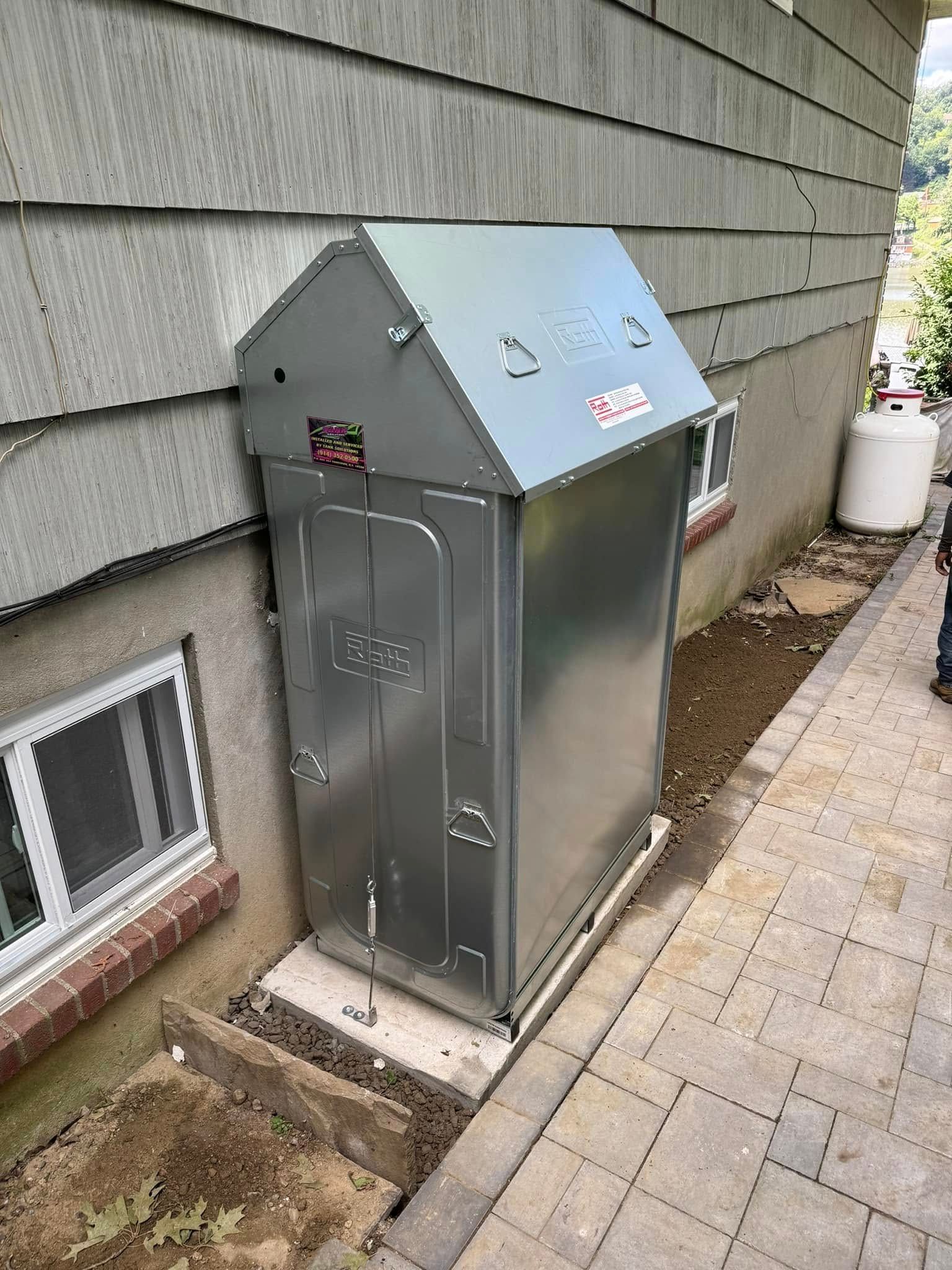 A tall, silver metal storage cabinet sits on a stone base against the side of a house next to a window.
