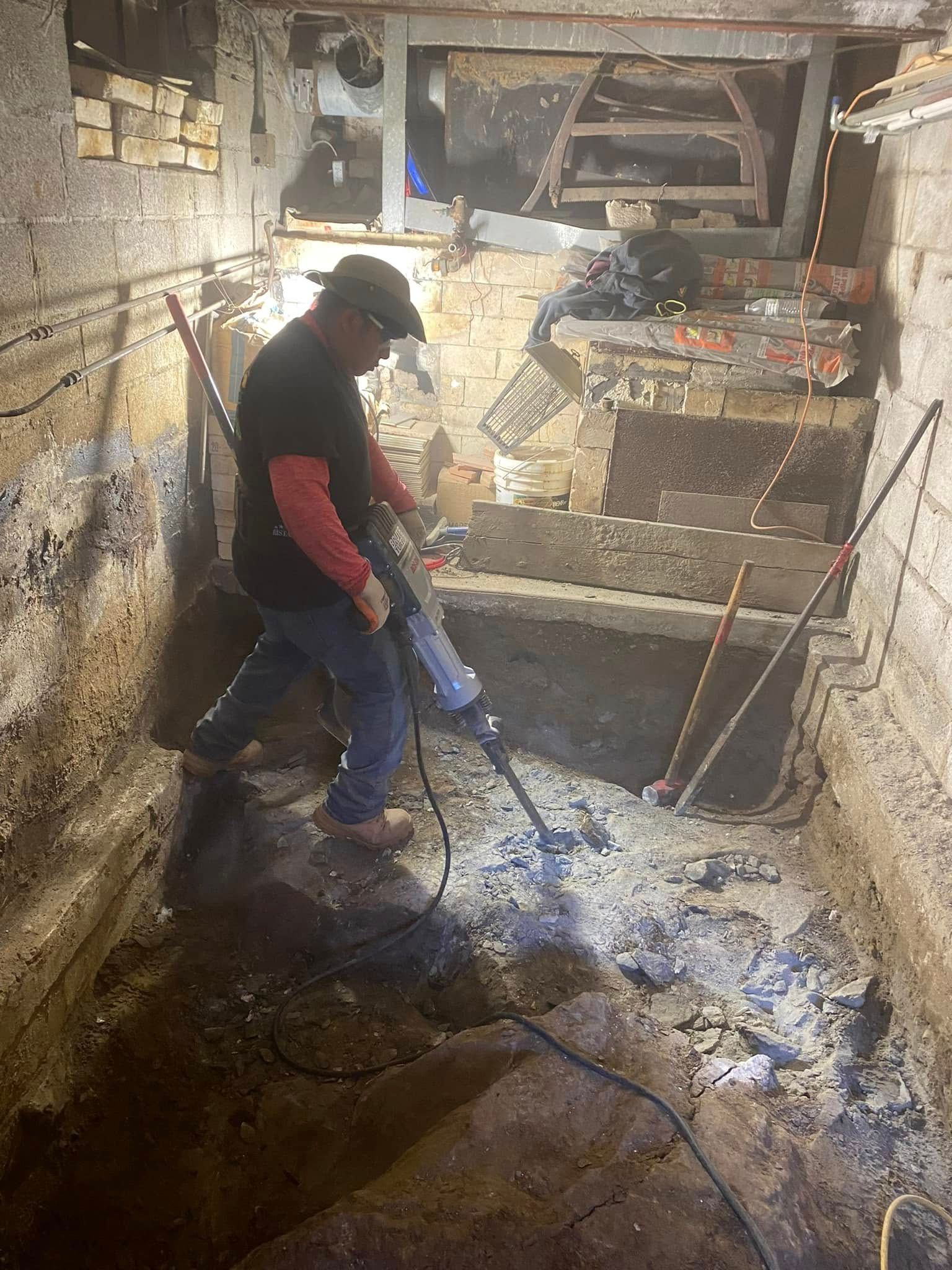 A worker in a basement uses a jackhammer to break up a concrete floor.